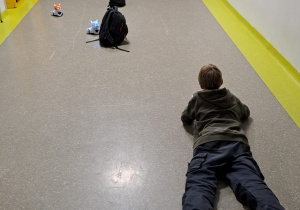 Na szkolnym korytarzu leży chłopiec na brzuchu, rozłożone są plecaki. Między plecakami widać roboty sterowane przez uczniów. W głębi korytarza widać jeszcze dwóch chłopców.