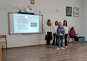 Na zdjęciu widać cztery uczennice śpiewające kolędę w języku niemieckim. Na tablicy multimedialnej jest wyświetlony tekst kolędy. Po lewej stronie siedzi uczennica prowadząca koncert.