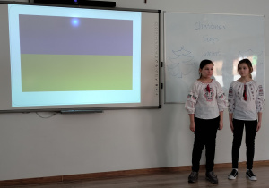 Na tablicy multimedialnej widać flagę Ukrainy. Po prawej stronie stoją dwie dziewczynki ubrane w koszule z haftem z Ukrainy.