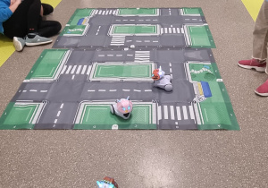 Na zdjęciu korytarz szkolny. Na korytarzu rozłożona jest makieta miasteczka drogowego. Z przodu widać jadącego robota, po lewej stronie zdjęcia siedzi uczeń i steruje robotem.