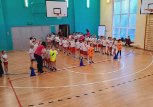Zdjęcie zrobione z góry. Widok na salę gimnastyczną. Widać dzieci ustawione w trzy rzędy przygotowane do rozpoczęcia konkurencji sportowej. Uczniowie ubrani są w biało-czerwone stroje sportowe.