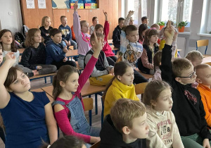 Na zdjęciu dzieci siedzą w klasie, biorą udział w spotkaniu. Niektóre dzieci mają uniesioną rękę do góry, chcą odpowiedzieć na zadane pytanie przez prowadzącego.