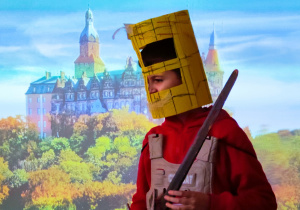 Na zdjęciu uczeń przebrany za rycerza, na głowie ma zrobiony własnoręcznie hełm w kolorze żółtym, założony czerwony płaszcz, trzyma w ręku miecz. W tle na tablicy multimedialnej wyświetlony jest zamek