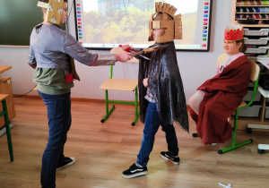 W sali szkolnej dwójka uczniów odgrywa scenę pojedynku rycerskiego. Na głowach mają hełmy, w rękach trzymają miecze.