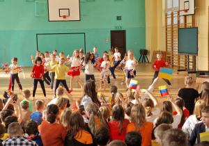 Na sali gimnastycznej dzieci oglądają tańczących kolegów i koleżanki. Wszystkie dzieci ubrane są kolorowe. Niektóre trzymają w rękach flagi Wenezueli i Ukrainy.