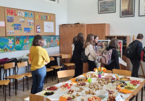 Na zdjęciu sala szkolna. Na środku stoi stół z typowymi przysmakami holenderskimi. Na ścianie widać dekorację oraz rysunki wiatraków. Dookoła stoją dzieci i oglądają wystawę poświęconą Holandii.