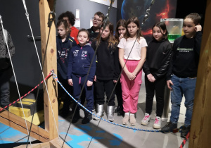 Grupa dzieci stoi przed dużym urządzeniem - maszyną "perpetuum mobilie".Widać drewnianą konstrukcję oraz duże zielone kule.