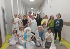 Zdjęcie grupowe uczniów na korytarzu szkolnym. Są przebrani za greckich bogów. W ubiorze dominuje kolor biały. Niektórzy w rękach trzymają atrybuty greckich bogów.