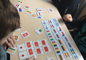 Przy stoliku siedzą dzieci. Na stole rozłożone są kartoniki z flagami państw z całego świata.