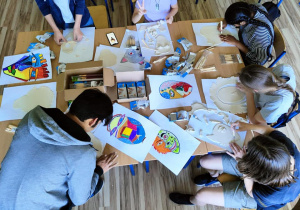 Uczniowie siedzą wokół stołu. Na stole rozłożone prace plastyczne. Na kartkach narysowane są projekty masek. Widać jak dzieci wycinają kształt maski z masy plastycznej. Na stole stoją również kartony z przyborami plastycznymi.