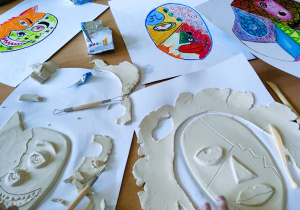 Na stole rozłożone prace plastyczne. Na kartkach narysowane są projekty masek. Widać jak dzieci wycinają kształt maski z masy plastycznej.