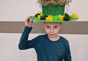 Na zdjęciu chłopiec ubrany w zieloną bluzkę oraz z kapeluszem na głowie. Kapelusz jest wiosennie ozdobiony. Na czubku jest gniazdo, a w gnieździe siedzi bocian. Chłopiec przytrzymuje kapelusz prawą ręką.