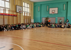 Na sali gimnastycznej dookoła siedzą dzieci. Widać, że niektóre z nich mają na sobie zielone elementu ubioru.