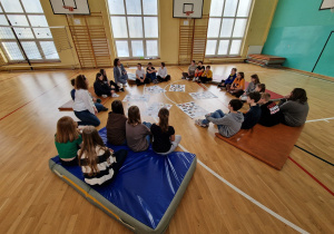 Na sali gimnastycznej uczniowie siedzą na materacach rozłożonych w okrąg. Przed nimi znajdują się arkusze białego papieru z obrazkami.