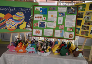 Na zdjęciu stoją trzy tablice z informacjami o świętach, na stole stoi wystawa kolorowych zajączków wielkanocnych wykonanych z papieru.