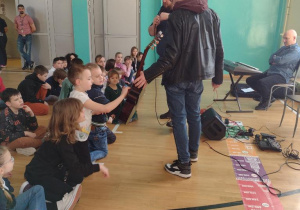 Na sali gimnastycznej po lewej stronie kadru siedzą uczniowie. Po prawej stronie stoją muzycy z gitarą. Podchodzą do dzieci, aby mogły z bliska obejrzeć gitarę.