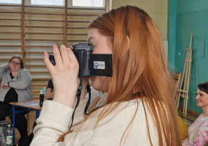 Uczennica trzyma w rękach aparat, robi zdjęcia podczas spotkania projektowego. Na aparacie widać logo programu "Laboratoria Przyszłości"