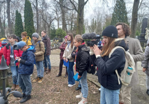 Na cmentarzu widać grupę stojących dzieci. Wśród nich po prawej stronie widać uczennicę z kamerą