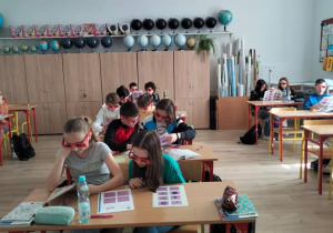 Sala szkolna. Uczniowie siedzą w ławkach. Na ławce rozłożone kartki z figurami, które można oglądać w okularach 3D. Uczniowie takie okulary mają założone.