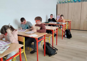 Sala szkolna. Uczniowie siedzą w ławkach. Na ławce rozłożone kartki z figurami, które można oglądać w okularach 3D. Uczniowie takie okulary mają założone.