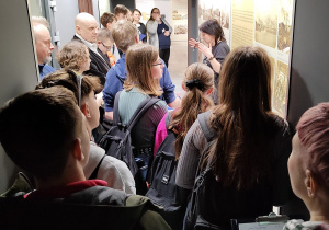 Grupa uczniów stoi w korytarzu muzeum. Widać przewodnika opowiadającego o ekspozycji.
