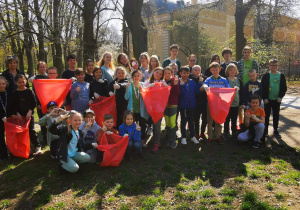 Zdjęcie grupowe uczniów. Stoją na trawie w parku. W rękach trzymają czerwone worki na śmieci.