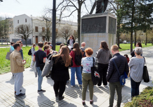 Uczniowie stoją na Placu Katedralnym przy pomniku księdza Ignacego Skorupki i słuchają opowieści nauczycielki historii.