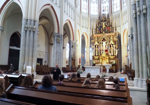 Uczniowie siędzą w ławkach ustawionych w katedrze, przodem do ołtarza głównego. Ksiądz proboszcz opowiada historię parafii.