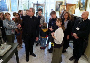 Grupa uczniów stoi w sali muzeum katedralnego. Widać eksponaty umieszczone w gablotach. Ksiądz proboszcz opowiada historię zebranych przedmiotów muzealnych.