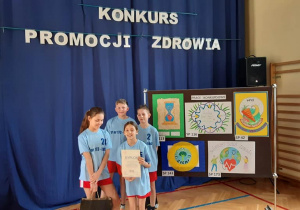 Czworo dzieci ubranych w niebieskie koszulki stoi obok tablicy z pracami konkursowymi. W tle wisi niebieska zasłona z napisem "Konkurs promocji zdrowia".