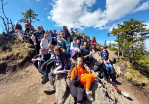 Zdjęcie grupowe uczniów na górze. Uczniowie siedzą na kamieniach. Widać drzewa oraz roślinność górską.