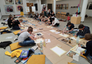 Na podłodze wokół rozłożonego szarego papieru siedzą uczniowie. Na papierze rozłożone są kartony i obrazy. Uczniowie siedzą w sali Miejskiej Galerii Sztuki