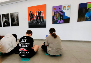 Troje uczniów siedzi tyłem do oglądającego zdjęcie, a przodem do ściany z obrazami.