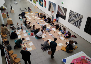 Widok z góry na salę Miejskiej Galerii Sztuki. Widać uczniów siedzących wokół szarego papieru, na którym rozłożone są kartony i przybory malarskie. Na ścianach wiszą obrazy.