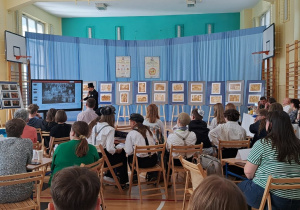 Zdjęcie ogólne sali gimnastycznej. Na sali siedzą zgromadzeni uczestnicy projektu. Widać rozstawione zdjęcia i prace konkursowe, tablicę multimedialną i prelegenta opowiadającego historię Łodzi.