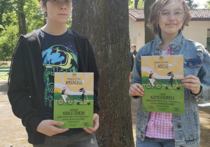 Dwoje uczniów stoi obok drzewa. Trzymają w rękach dyplomy za udział w konkursie plastycznym.