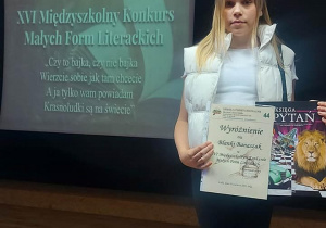 Na zdjęciu uczennica stoi obok tablicy multimedialnej z wyświetloną nazwą konkursu. Blanka w ręku trzyma dyplom oraz nagrodę rzeczową.