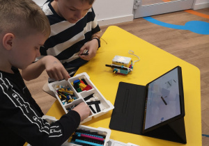 Dwóch uczniów siedzi przy żółtej ławce. Na stole stoi tablet, pudełko z klockami, kartki oraz mały robot.