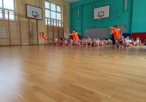 Sala gimnastyczna. Widać trzy drużyny. Dzieci siedzą jedno za drugim. Troje dzieci w pomarańczowych koszulkach biegnie z piłkami w odwróconym pachołku.