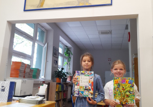 Na zdjęciu w bibliotece szkolnej stoją dwie dziewczynki i trzymają w rękach książki Juliana Tuwima