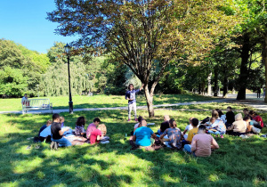 W parku na trawie siedzą uczniowie, słuchają nauczyciela prowadzącego zajęcia.