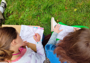 Na zdjęciu widać dwie dziewczynki rysujące obrazki. Na obrazkach widać drzewo. Dziewczynki siedzą na trawie.