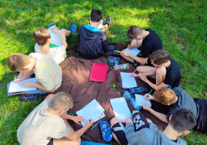Na zdjęciu widać grupę uczniów siedzących na trawie i wykonujących rysunki.