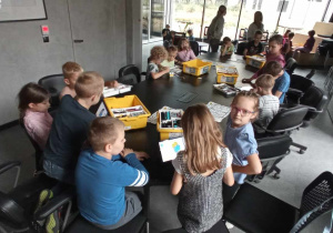 Na krzesłach dookoła owalnego stołu siedzą dzieci. Przy każdym dziecku stoi na stole żółte pudełko, w którym znajdują się zestawy do zajęć z robotyki.