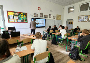 W sali szkolnej w ławkach siedzą uczniowie, słuchają prelekcji prowadzącego.