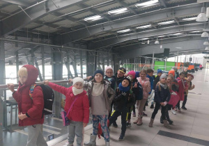 Grupa dzieci stoi przy barierkach w hali portu lotniczego w Łodzi.