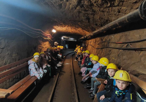 W korytarzu kopalni po obu stronach ścian na ławkach siedzą dzieci w żółtych kaskach na głowach. Na ziemi widać tor kolejowy.