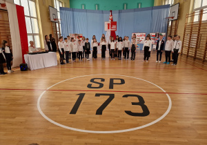 Na sali gimnastycznej stoją uczniowie śpiewający w chórze. Z tyłu widać dekorację. Z przodu na podłodze widnieje napis SP 173.