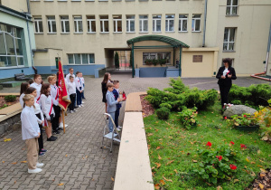 Na dziedzińcu szkolnym grupa uczniów stoi przed trawnikiem, na którym stoi pani dyrektor. Jest to moment złożenia kwiatów pod dębem zasadzonym z okazji 100-lecia niepodległości.