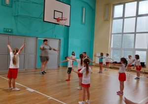 Na sali gimnastycznej stoją dzieci ubrane w biało-czerwone stroje sportowe. Osoba prowadząca zajęcia pokazuje ćwiczenia.